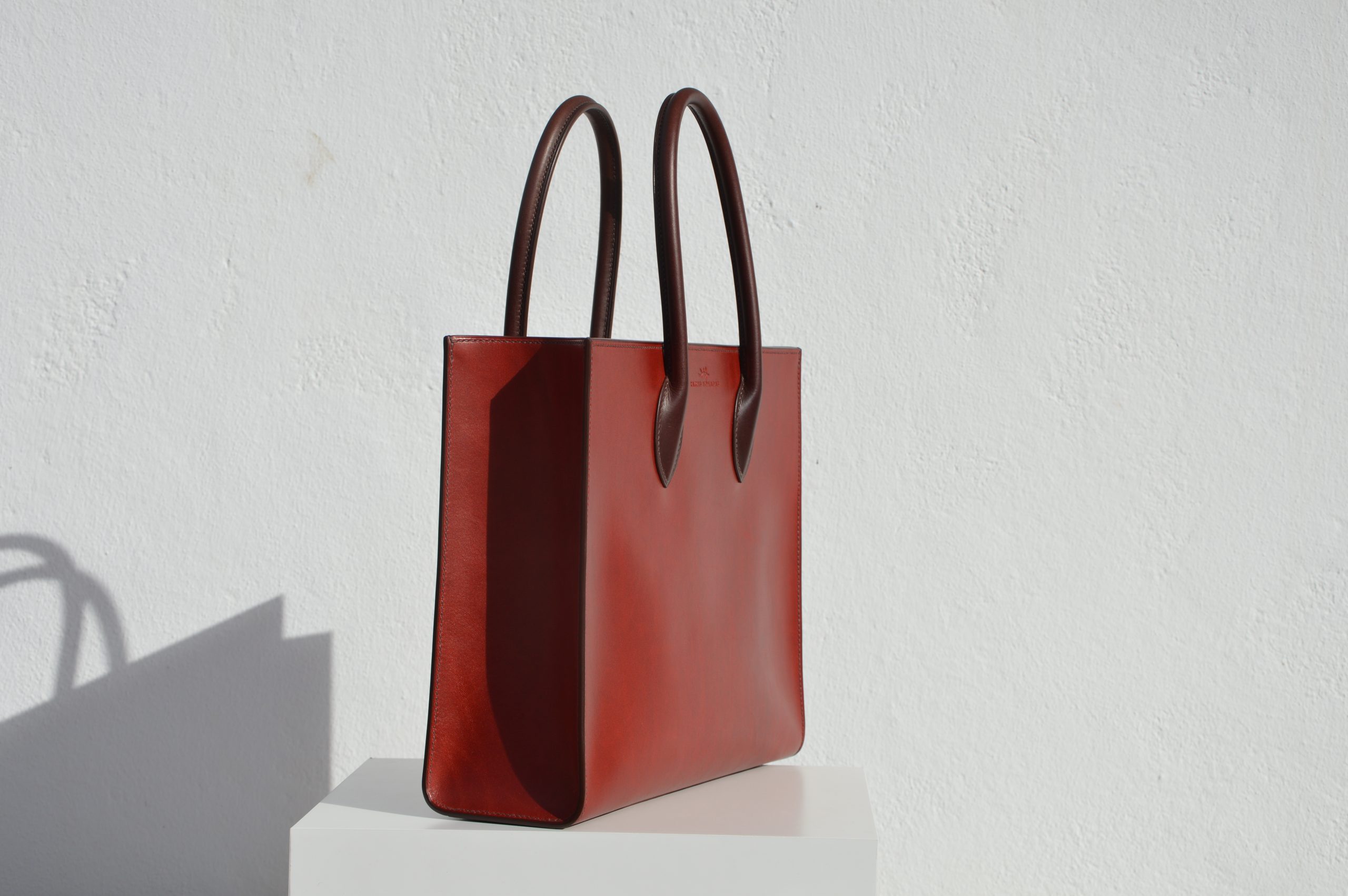 "Tote Bag con cremallera disponible por €1,400.00. Este clásico bolso combina belleza atemporal y líneas armónicas. Creado con el máximo cuidado, está confeccionado con la mejor piel de curtido vegetal española, hilos de lino y cosido completamente a mano. El bolso artesanal presenta un elegante color cereza con asas color tabaco, y está forrado con piel de cabra francesa. Cuenta con un bolsillo interior sin cremallera y otro con cremallera, además de un cierre exterior. Cada detalle de este bolso refleja la artesanía y la calidad de su fabricación, 100% cosido a mano."