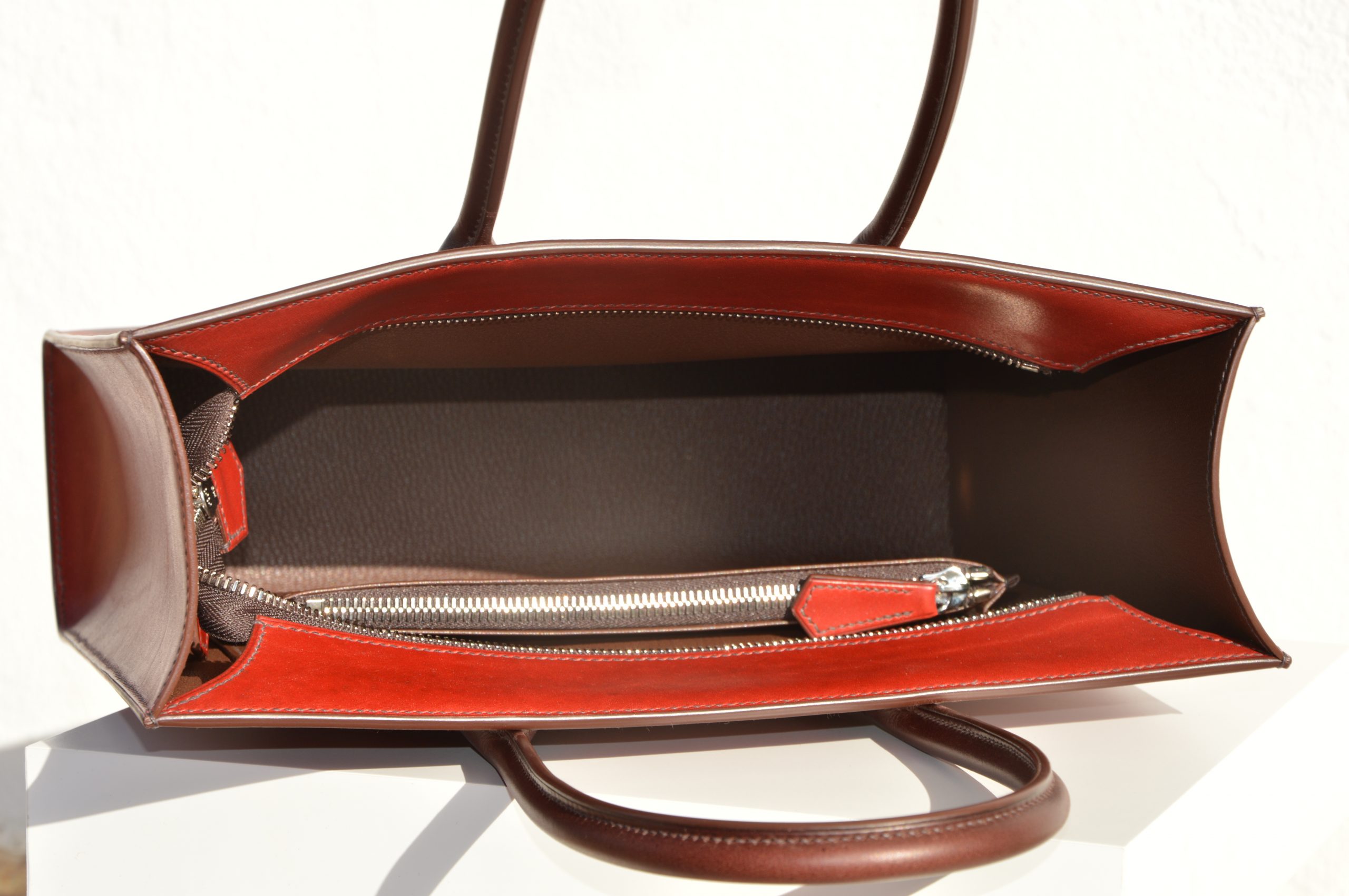 "Tote Bag con cremallera disponible por €1,400.00. Este clásico bolso combina belleza atemporal y líneas armónicas. Creado con el máximo cuidado, está confeccionado con la mejor piel de curtido vegetal española, hilos de lino y cosido completamente a mano. El bolso artesanal presenta un elegante color cereza con asas color tabaco, y está forrado con piel de cabra francesa. Cuenta con un bolsillo interior sin cremallera y otro con cremallera, además de un cierre exterior. Cada detalle de este bolso refleja la artesanía y la calidad de su fabricación, 100% cosido a mano."
