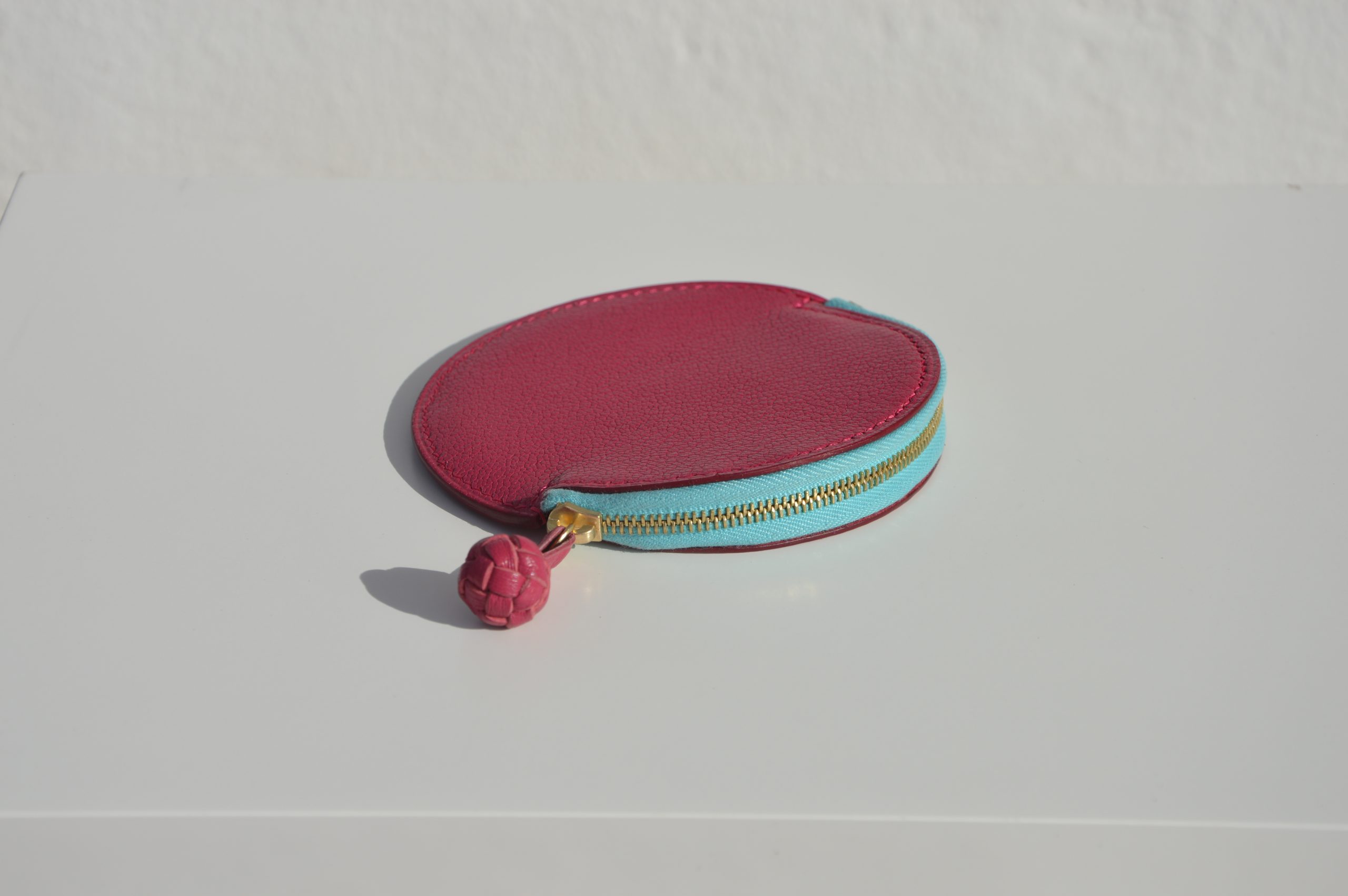 "Monedero Redondo disponible por €115.00. Elaborado con piel de cabra de origen francés, con detalle de tirador trenzado. Cosido completamente a mano con hilos de lino y equipado con una cremallera de la mejor calidad. Personalizable en colores de piel y hilos. Una pieza artesanal que combina estilo y funcionalidad."