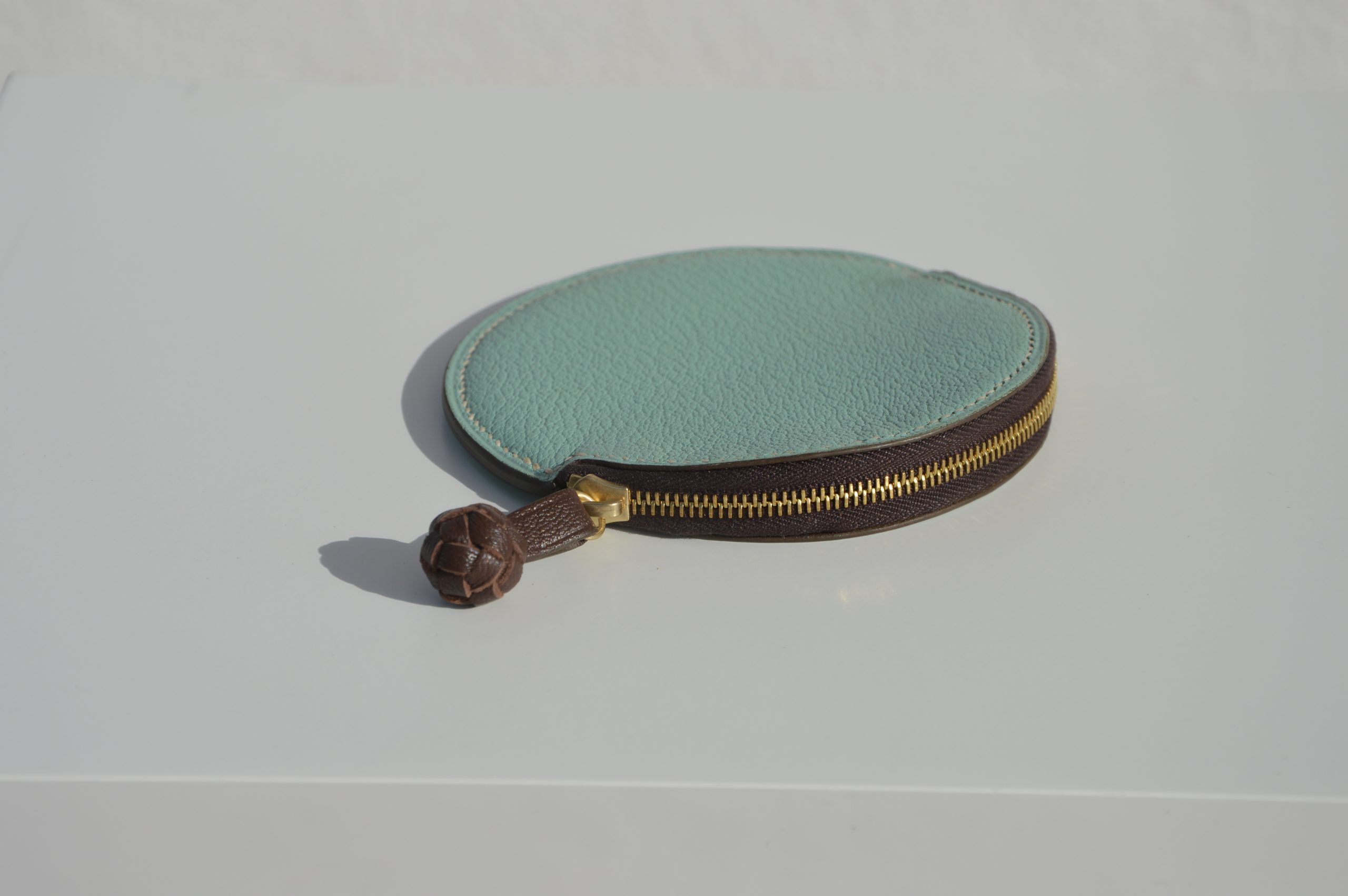 "Monedero Redondo disponible por €115.00. Elaborado con piel de cabra de origen francés, con detalle de tirador trenzado. Cosido completamente a mano con hilos de lino y equipado con una cremallera de la mejor calidad. Personalizable en colores de piel y hilos. Una pieza artesanal que combina estilo y funcionalidad."