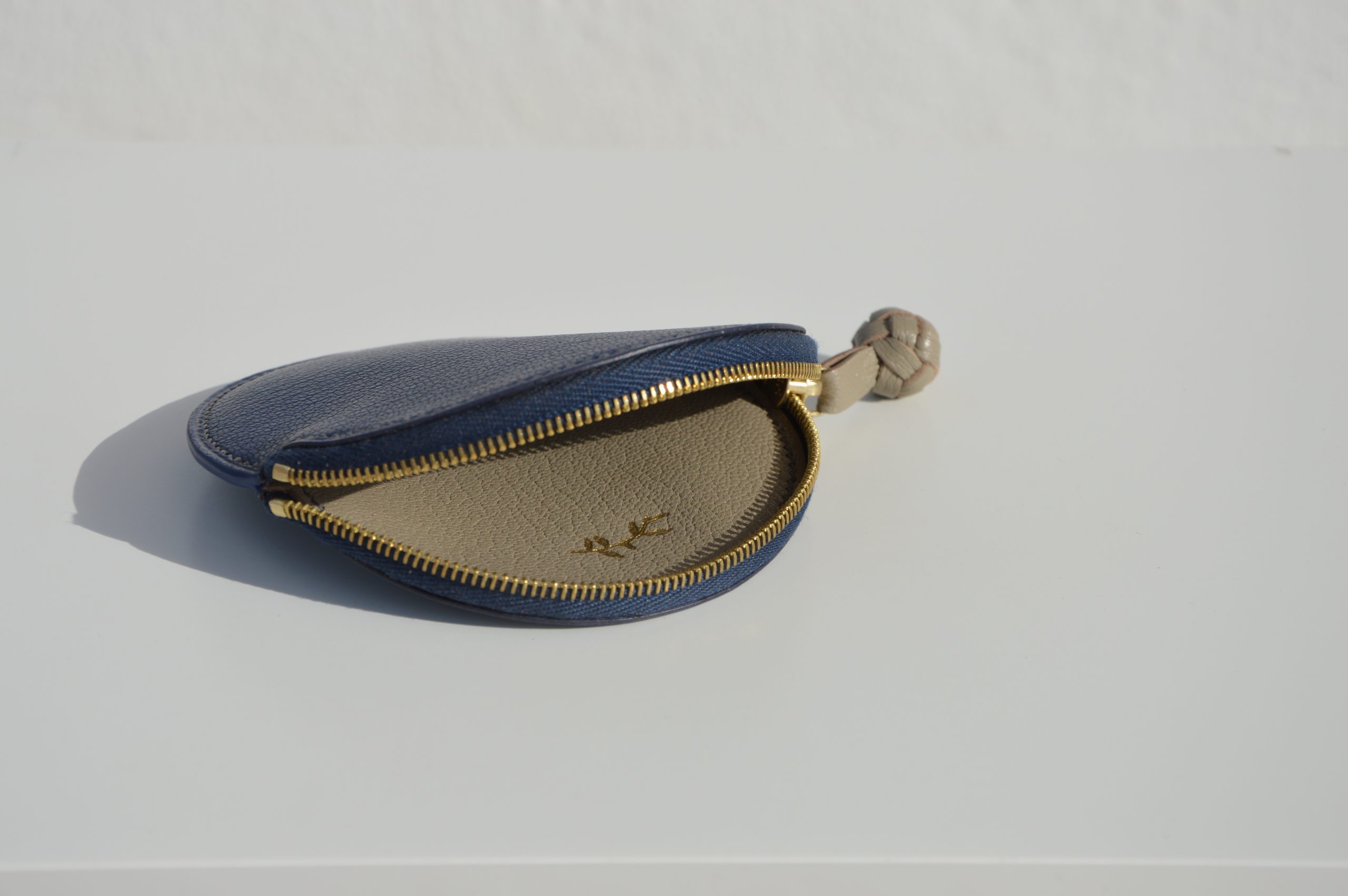 "Monedero Redondo disponible por €115.00. Elaborado con piel de cabra de origen francés, con detalle de tirador trenzado. Cosido completamente a mano con hilos de lino y equipado con una cremallera de la mejor calidad. Personalizable en colores de piel y hilos. Una pieza artesanal que combina estilo y funcionalidad."