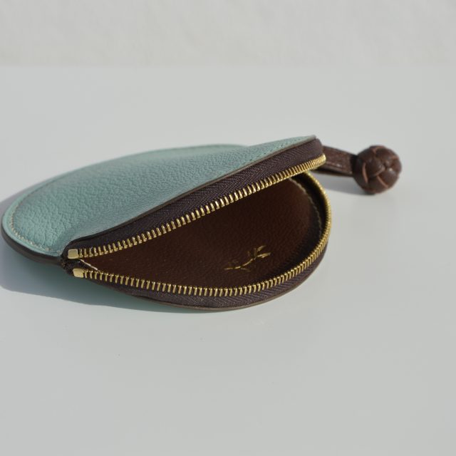 "Monedero Redondo disponible por €115.00. Elaborado con piel de cabra de origen francés, con detalle de tirador trenzado. Cosido completamente a mano con hilos de lino y equipado con una cremallera de la mejor calidad. Personalizable en colores de piel y hilos. Una pieza artesanal que combina estilo y funcionalidad."