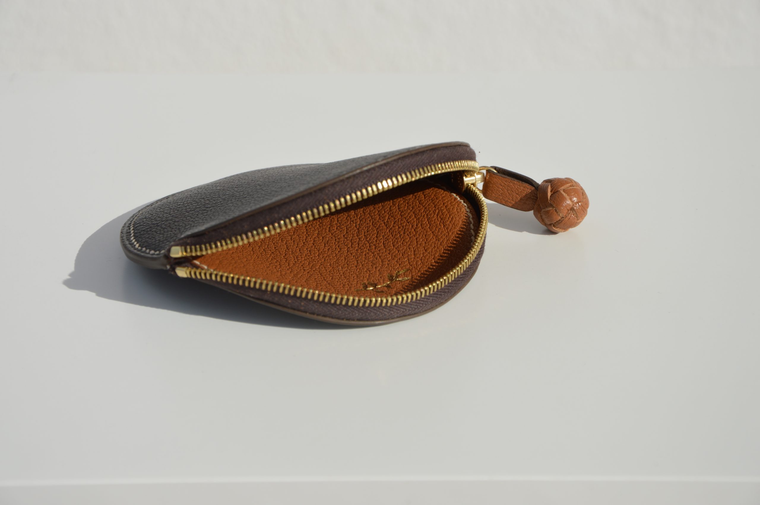 "Monedero Redondo disponible por €115.00. Elaborado con piel de cabra de origen francés, con detalle de tirador trenzado. Cosido completamente a mano con hilos de lino y equipado con una cremallera de la mejor calidad. Personalizable en colores de piel y hilos. Una pieza artesanal que combina estilo y funcionalidad."