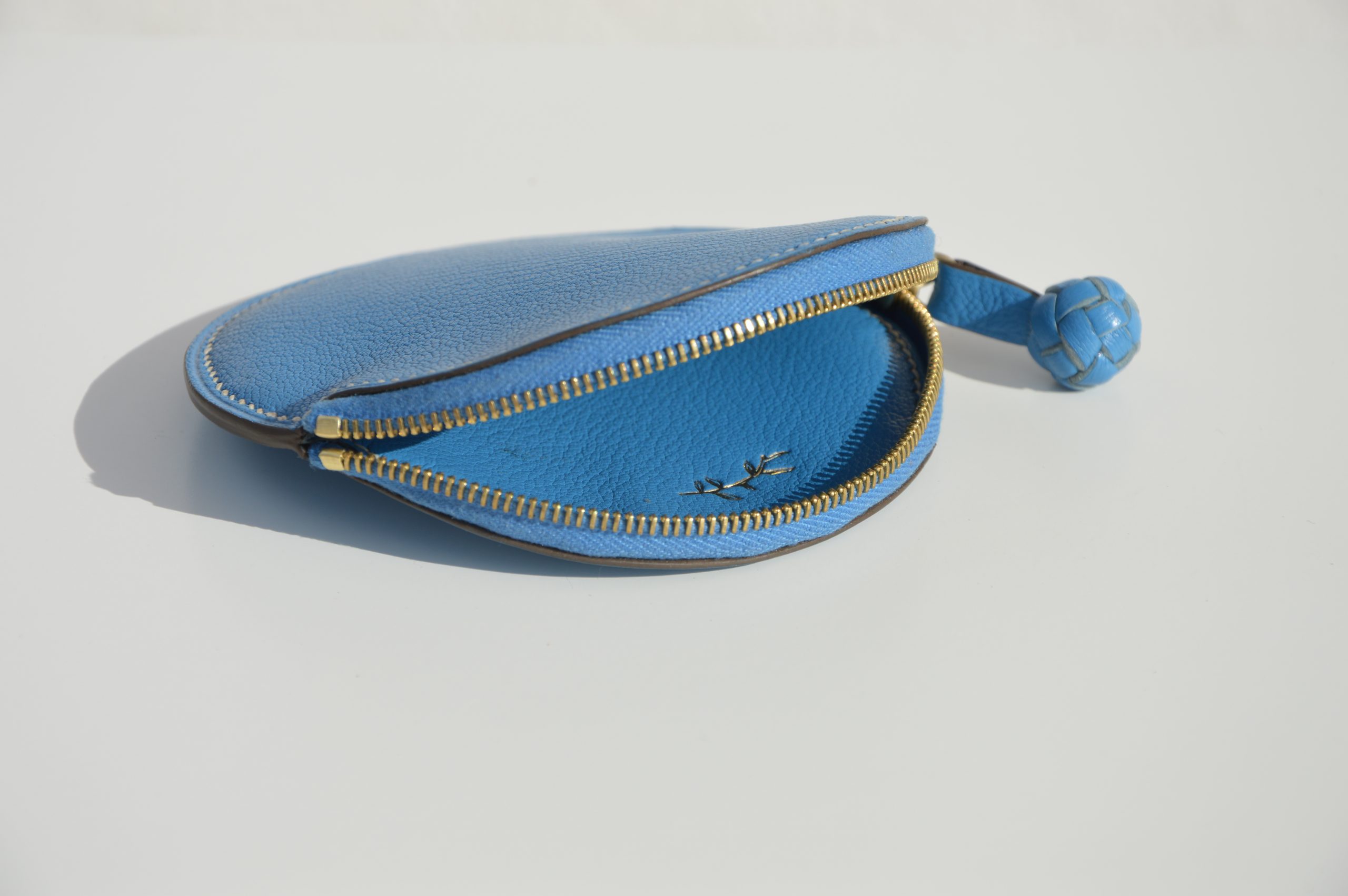 "Monedero Redondo disponible por €115.00. Elaborado con piel de cabra de origen francés, con detalle de tirador trenzado. Cosido completamente a mano con hilos de lino y equipado con una cremallera de la mejor calidad. Personalizable en colores de piel y hilos. Una pieza artesanal que combina estilo y funcionalidad."