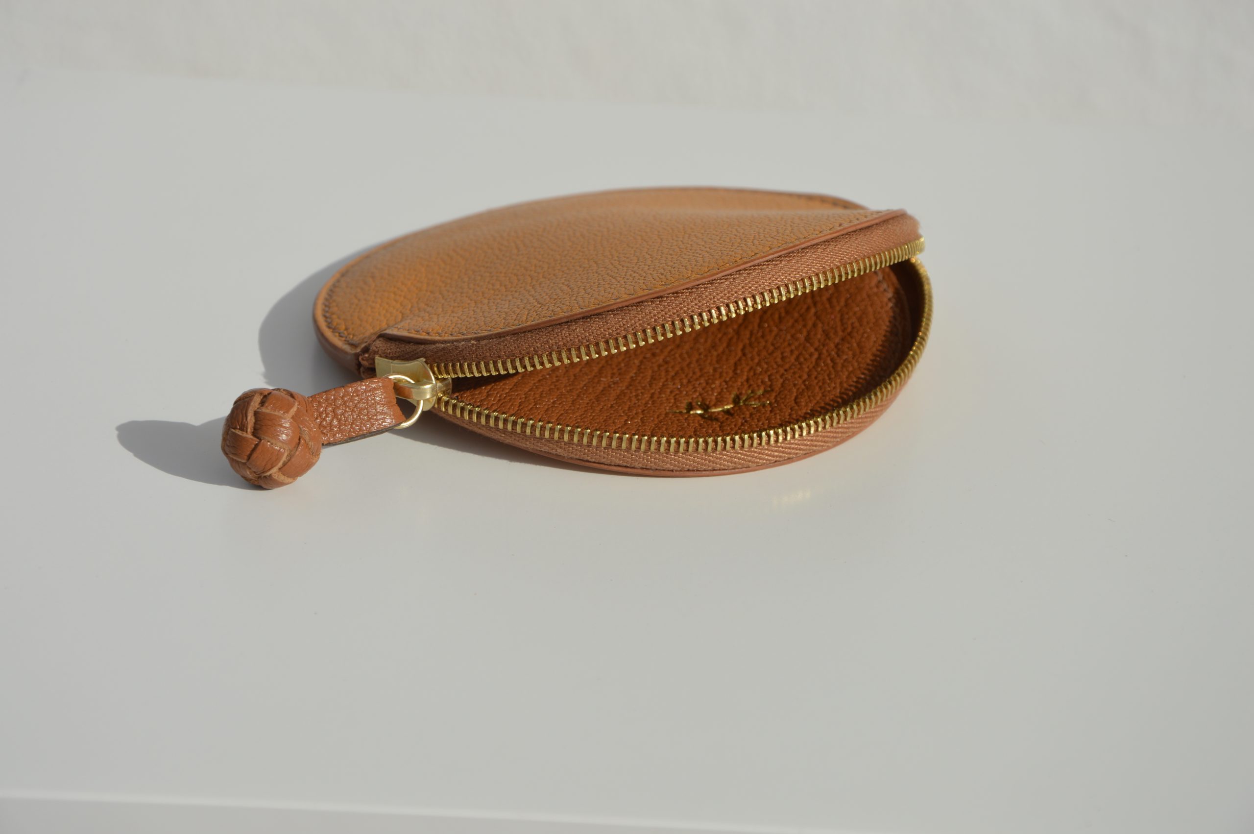 "Monedero Redondo disponible por €115.00. Elaborado con piel de cabra de origen francés, con detalle de tirador trenzado. Cosido completamente a mano con hilos de lino y equipado con una cremallera de la mejor calidad. Personalizable en colores de piel y hilos. Una pieza artesanal que combina estilo y funcionalidad."