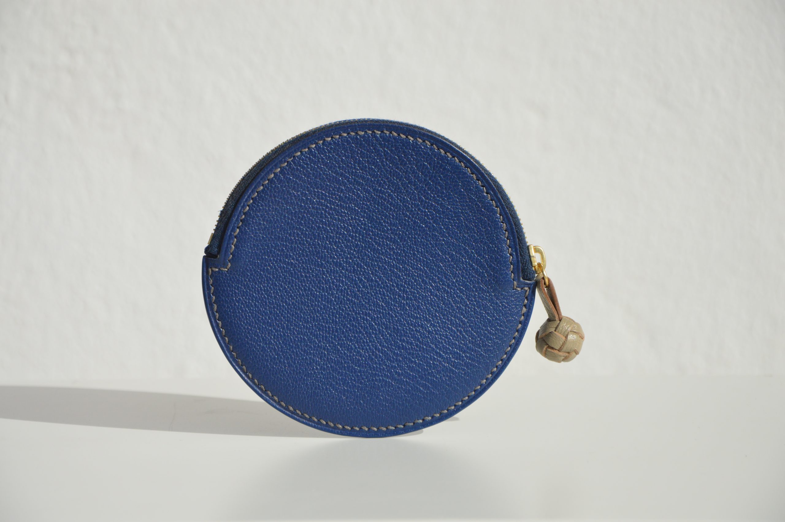 "Monedero Redondo disponible por €115.00. Elaborado con piel de cabra de origen francés, con detalle de tirador trenzado. Cosido completamente a mano con hilos de lino y equipado con una cremallera de la mejor calidad. Personalizable en colores de piel y hilos. Una pieza artesanal que combina estilo y funcionalidad."