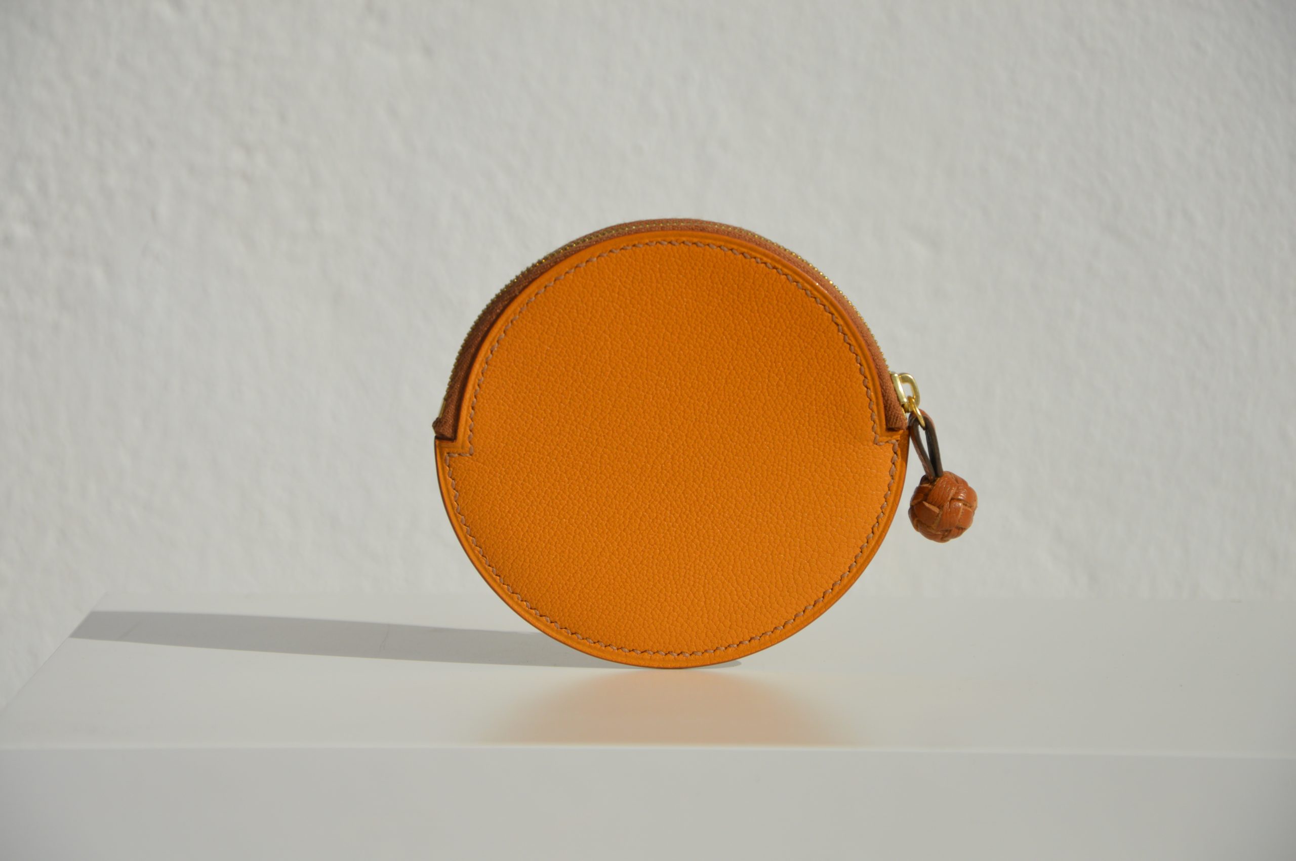 "Monedero Redondo disponible por €115.00. Elaborado con piel de cabra de origen francés, con detalle de tirador trenzado. Cosido completamente a mano con hilos de lino y equipado con una cremallera de la mejor calidad. Personalizable en colores de piel y hilos. Una pieza artesanal que combina estilo y funcionalidad."