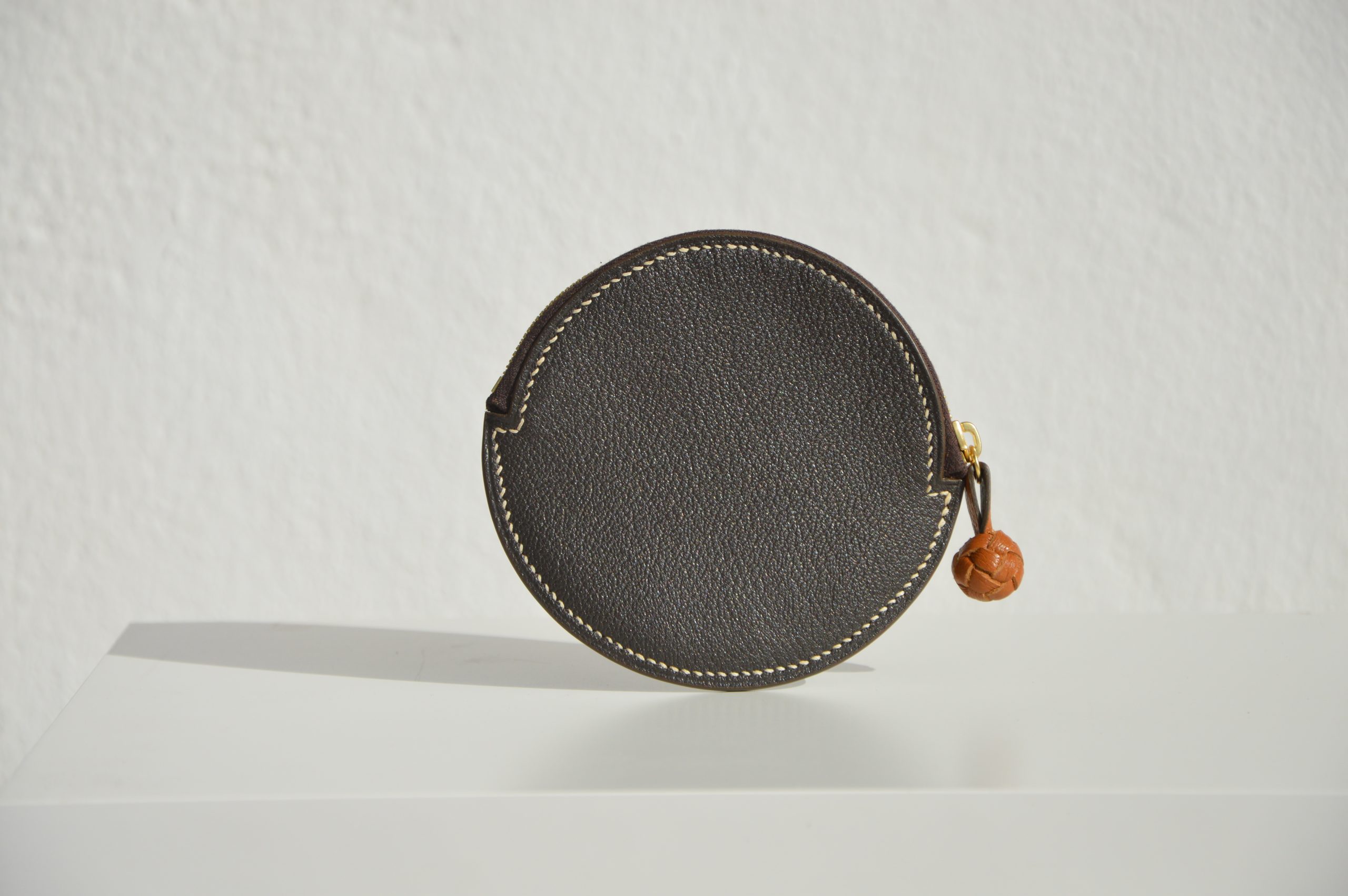 "Monedero Redondo disponible por €115.00. Elaborado con piel de cabra de origen francés, con detalle de tirador trenzado. Cosido completamente a mano con hilos de lino y equipado con una cremallera de la mejor calidad. Personalizable en colores de piel y hilos. Una pieza artesanal que combina estilo y funcionalidad."