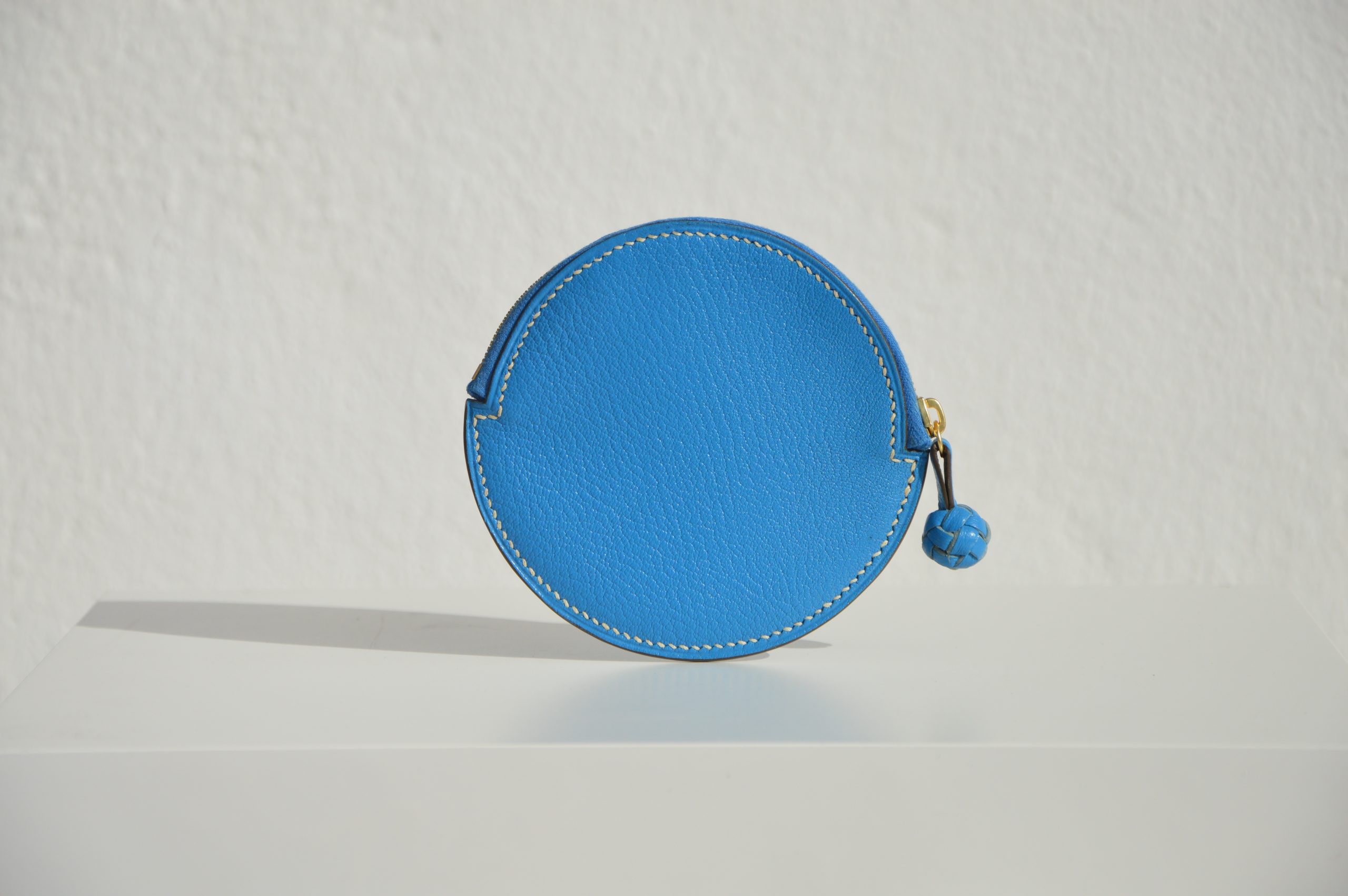 "Monedero Redondo disponible por €115.00. Elaborado con piel de cabra de origen francés, con detalle de tirador trenzado. Cosido completamente a mano con hilos de lino y equipado con una cremallera de la mejor calidad. Personalizable en colores de piel y hilos. Una pieza artesanal que combina estilo y funcionalidad."