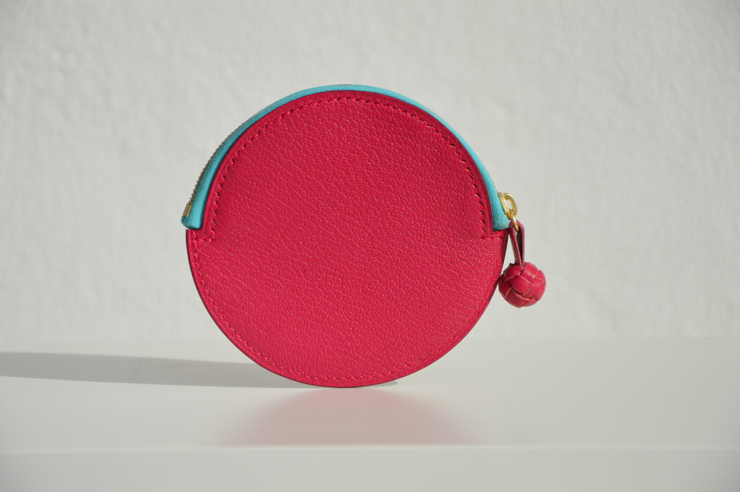 "Monedero Redondo disponible por €115.00. Elaborado con piel de cabra de origen francés, con detalle de tirador trenzado. Cosido completamente a mano con hilos de lino y equipado con una cremallera de la mejor calidad. Personalizable en colores de piel y hilos. Una pieza artesanal que combina estilo y funcionalidad."
